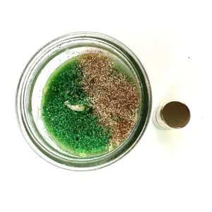 A jar of green and brown sand next to a bottle.